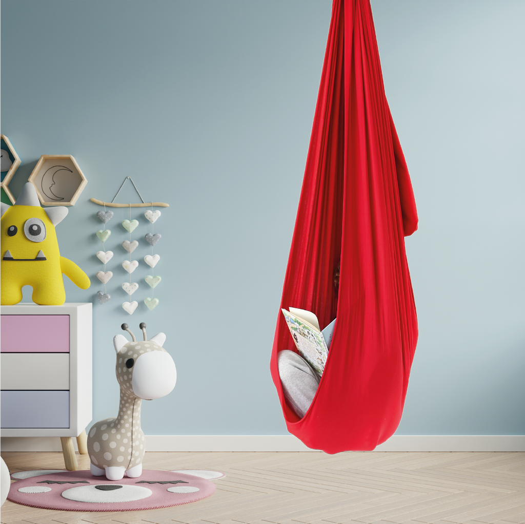An image for the post "What does a sensory swing do?" there is a red sensory swing with a child inside reading a book. The sensory swing is installed in the kids room there are toys and a dresser